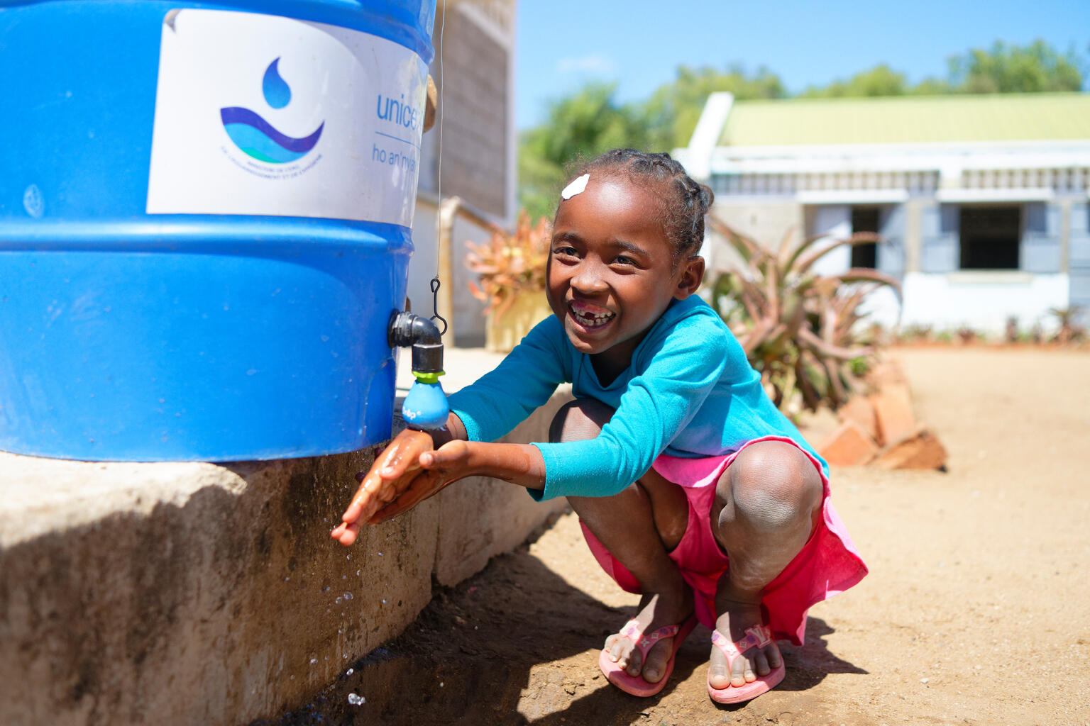 Ein lachendes Mädchen wäscht sich die Hände an einem blauen Wassertank mit UNICEF-Logo in einer sonnigen Außenumgebung.